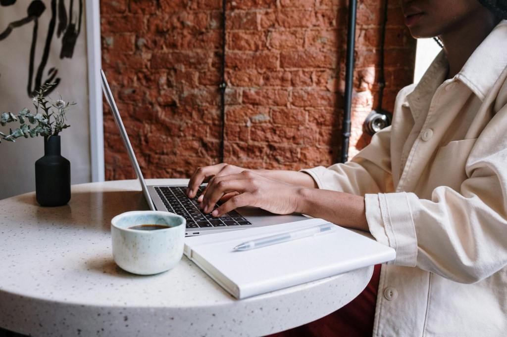 A person typing on a laptop at a round table, with a cup of coffee and a notepad nearby. The background features a brick wall and a small plant in a vase.