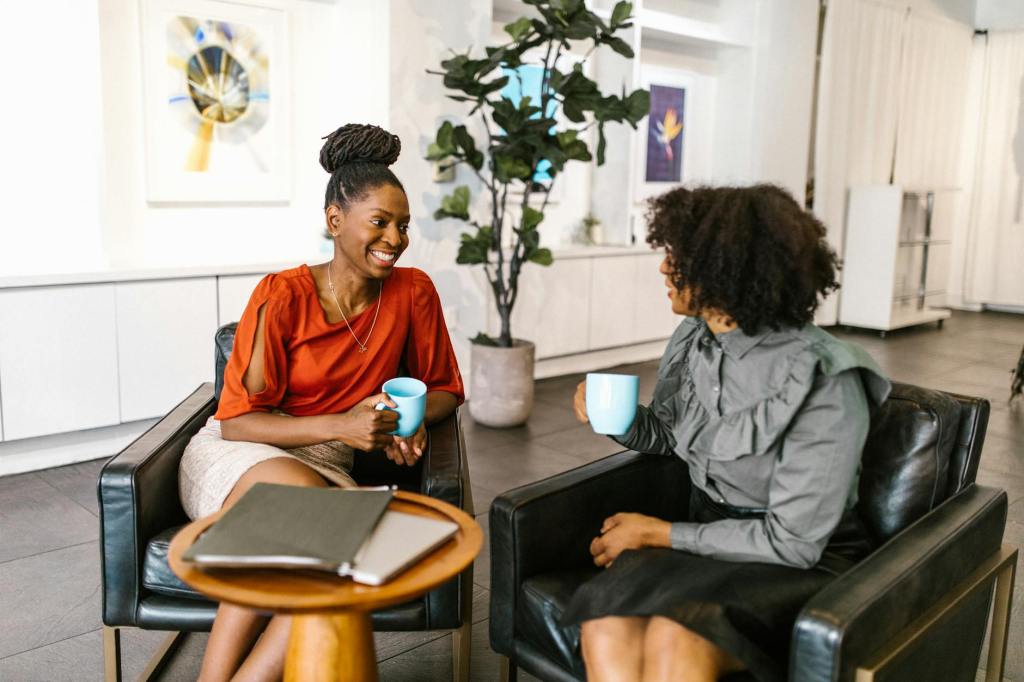 Two women sitting in a modern office setting, engaging in a conversation while holding coffee cups.